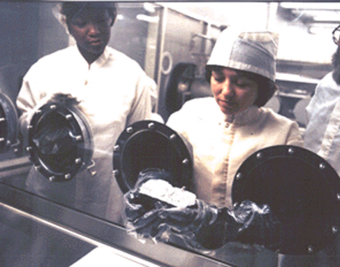 Photograph of Technicians Examining a Lunar Sample. Two scientists look at a Moon rock through the glass of a scientific “glove box”.