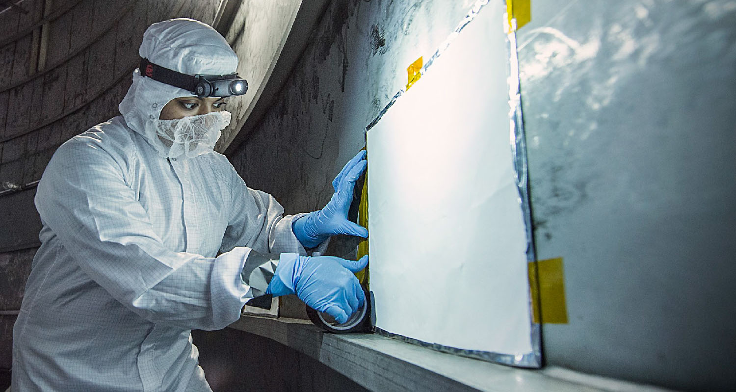 A person wearing a coverall and face mask attaches a rectangular panel to a wall.