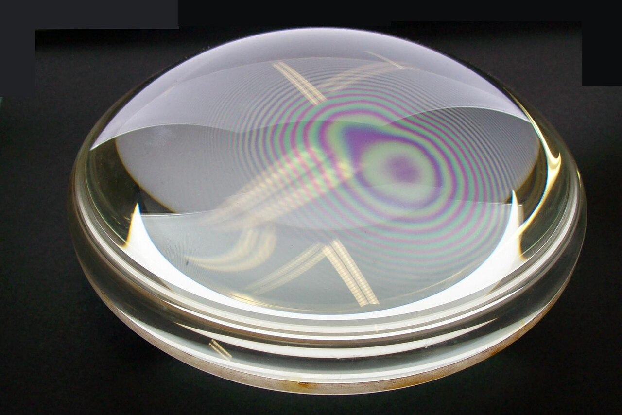 Newton’s Rings Interference Pattern - When a slightly curved glass lens is placed in contact with a flat glass plate, concentric circular fringes of alternating bright and dark regions appear around the contact point. These interference fringes, known as Newton’s rings, are created by the interference between light rays reflected from the bottom surface of the lens and the top surface of the glass plate.
The varying air gap thickness between the surfaces causes different path differences for the reflected rays, producing constructive interference (bright rings) where the path difference equals a whole number of wavelengths, and destructive interference (dark rings) where the path difference equals an odd number of half-wavelengths. This phenomenon puzzled Newton because it suggested wave-like behavior that was difficult to reconcile with his particle theory of light. The radius of each ring depends on the wavelength of light used, making Newton’s rings a precise method for measuring wavelengths and testing the flatness of optical surfaces.