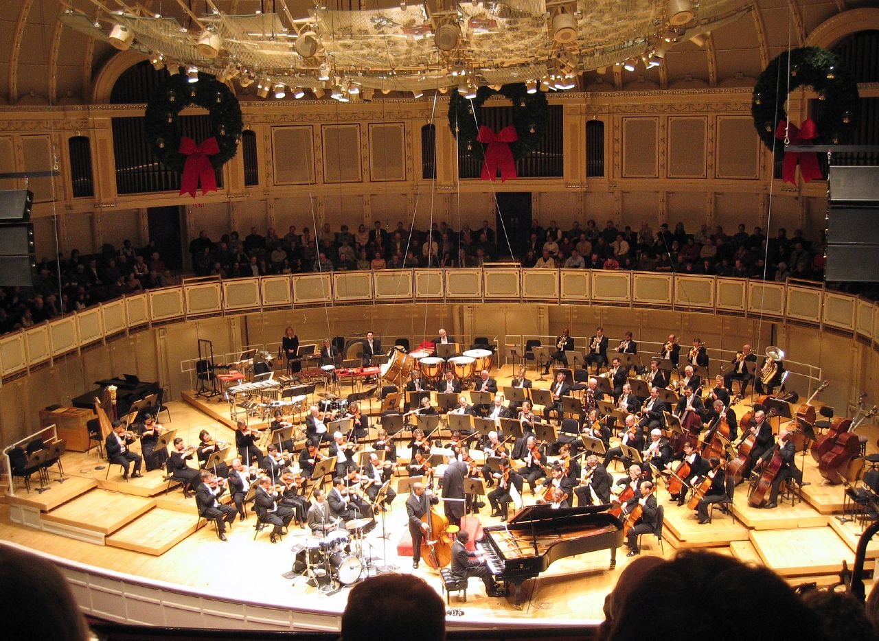 An orchestra performing on stage in a concert hall, seen from above, with the hall's balconies and a full audience surrounding it.